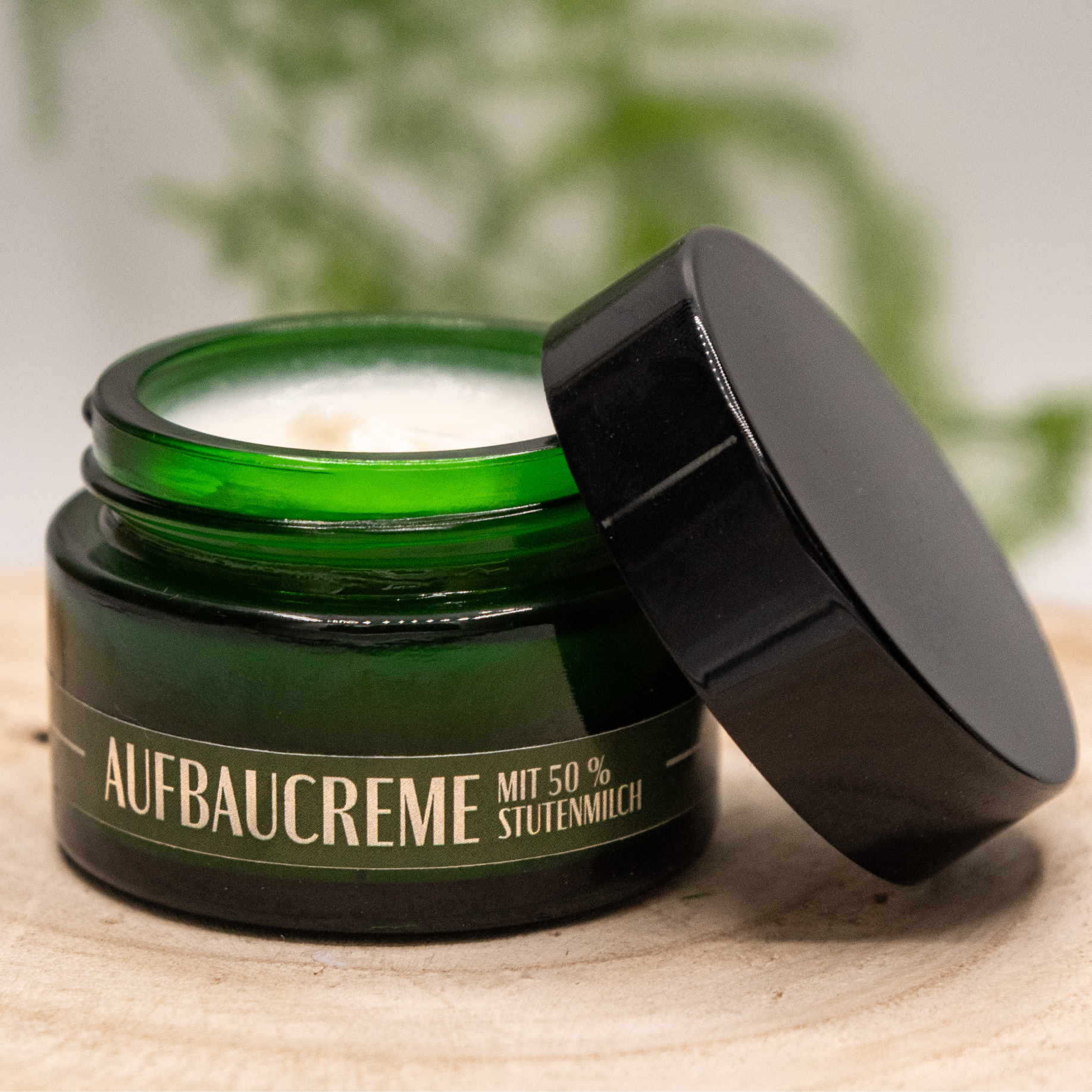 Eine geöffnete flaschengrüne Cremedose Aufbaucreme steht auf einer Baumscheibe, der schwarz glänzende Deckel liegt leicht schräg auf der Dose. Im Inneren sieht man die weiße Creme. "Aufbaucreme mit 50% Stutenmilch" steht in beider Schrift auf der Dose. Im Hintergrund ist unscharf ein grüner Zweig zu sehen.