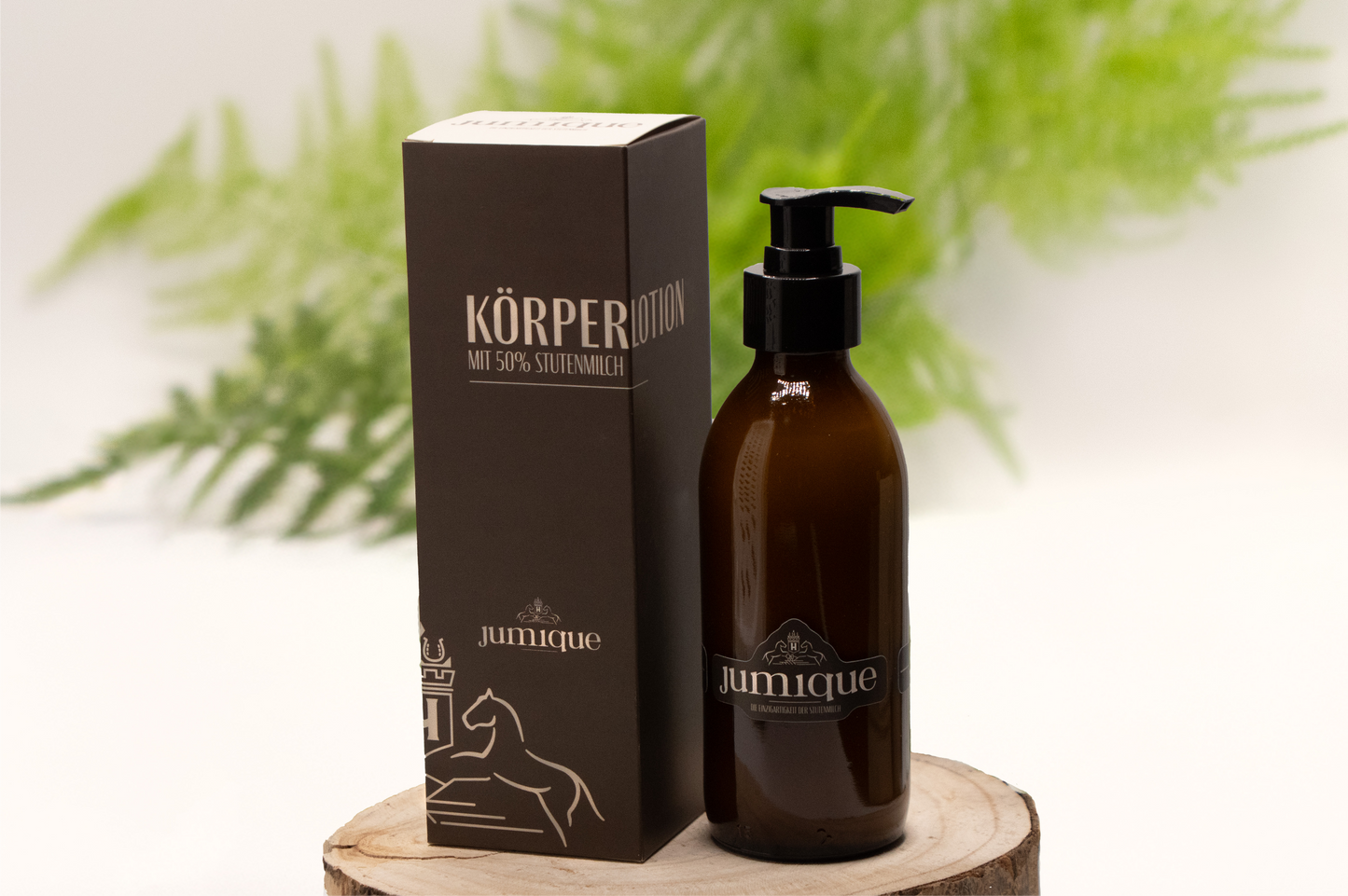Auf einer Baumscheibe steht eine braune Pumpflasche Körperlotion, darauf ist der Firmenname Jumique zu lesen. Das Logo- zwei Pferde die den Blick auf ein Wappen mit einem "H" im inneren gerichtet haben ist über dem Namen zu sehen. Daneben steht der Karton, in dem das Produkt verpackt wird. Er ist braun und trägt die beige Aufschrift "Körperlotion mit 50% Stutenmilch". Im überwiegend weißen Hintergrund ist unscharf ein grüner Zweig eines Farns zu sehen.