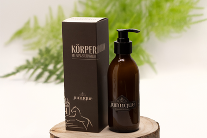 Auf einer Baumscheibe steht eine braune Pumpflasche Körperlotion, darauf ist der Firmenname Jumique zu lesen. Das Logo- zwei Pferde die den Blick auf ein Wappen mit einem "H" im inneren gerichtet haben ist über dem Namen zu sehen. Daneben steht der Karton, in dem das Produkt verpackt wird. Er ist braun und trägt die beige Aufschrift "Körperlotion mit 50% Stutenmilch". Im überwiegend weißen Hintergrund ist unscharf ein grüner Zweig eines Farns zu sehen.