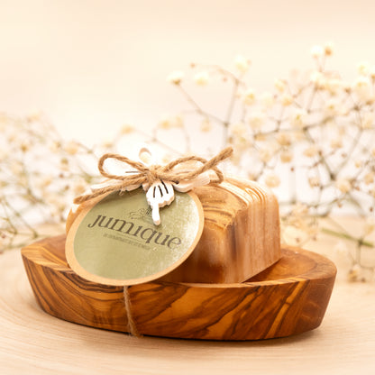Ein Seifenstück der Stutenmilchseife "Marquise" mit Hafer und Honig liegt in einer braun gemaserten Seifenschale aus Olivenholz. Darum ist mit einem braunen, geflochtenen Band ein grünes "Jumique" Schild und ein Schmetterling aus Holz gebunden. Die goldene und braune Maserung ist erkennbar. Das Bild hat einen warmen, goldenen Ton. Im unscharfen Hintergrund liegen dünne grüne Zweige mit zarten weißen Blüten.