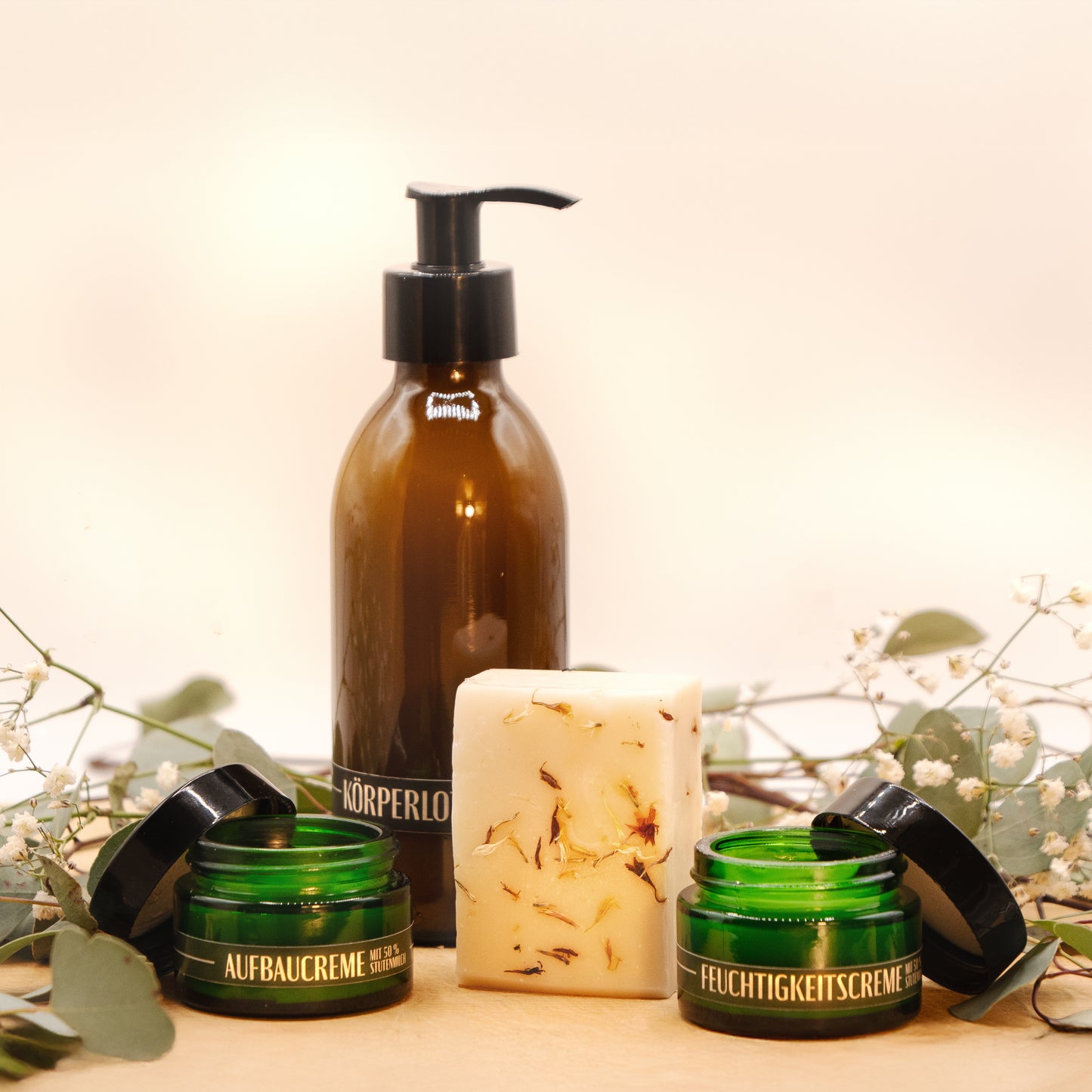 Ein Geschenkset steht auf einer Baumscheibe. Es besteht aus der Stutenmilchseife "Ouvertüre" mit Kardamom und Pfeffer, einer Körperlotion in einer braunen Pumpflasche sowie einer Aufbau- und einer Feuchtigkeitscreme in flaschengrünen Cremedosen. Das Seifenstück steht aufrecht in der Mitte, sodass die kleinen Kardamom-Stückchen sichtbar sind. Dahinter steht die Körperlotion Rechts und links stehen die Aufbau- und die Feuchtigkeitscreme. Zarte grüne Zweige mit weißen Blüten liegen im Hintergrund.