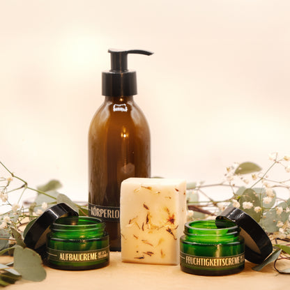 Ein Geschenkset steht auf einer Baumscheibe. Es besteht aus der Stutenmilchseife "Ouvertüre" mit Kardamom und Pfeffer, einer Körperlotion in einer braunen Pumpflasche sowie einer Aufbau- und einer Feuchtigkeitscreme in flaschengrünen Cremedosen. Das Seifenstück steht aufrecht in der Mitte, sodass die kleinen Kardamom-Stückchen sichtbar sind. Dahinter steht die Körperlotion Rechts und links stehen die Aufbau- und die Feuchtigkeitscreme. Zarte grüne Zweige mit weißen Blüten liegen im Hintergrund.