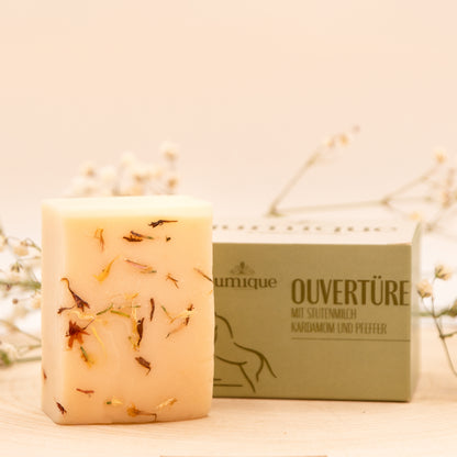 Ein Seifenstück der Stutenmilchseife "Ouvertüre" mit Kardamom und Pfeffer liegt neben dem dazugehörigen grünen Karton auf einer Baumscheibe. Es ist aufrecht hingestellt, sodass die kleinen Kardamonstücke erkennbar sind. Auf dem Karton steht in braun-grüner Schrift "Ouvertüre mit Stutenmilch, Kardamom und Pfeffer" rechts neben dem Jumique-Logo. Das Bild hat einen warmen, goldenen Ton. Im unscharfen Hintergrund liegen dünne grüne Zweige mit zarten weißen Blüten.