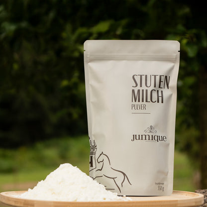 Eine weiße Packung steht auf einem Holzteller. In grau-grüner Schrift steht "Stutenmilch Pulver" auf der Vorderseite. Darunter steht der Firmenname Jumique. Das Logo- zwei Pferde die den Blick auf ein Wappen mit einem "H" im inneren gerichtet haben ist über dem Namen und am Rand der Verpackung zu sehen. Die Nettofüllmenge 350g ist ebenfalls auf der Packung angegeben. Neben der Packung liegt ein kleiner Haufen weißes Stutenmilch Pulver. Der Hintergrund ist ein unscharfes Grün der Bäume.