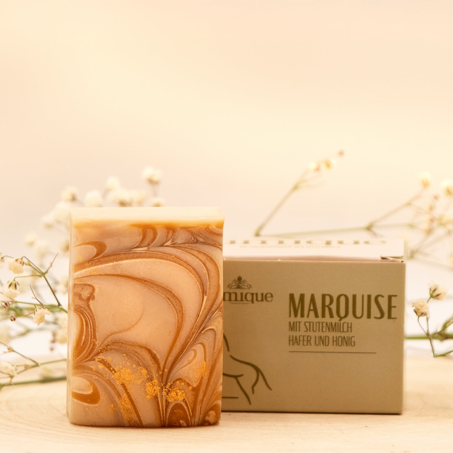 Ein Seifenstück der Stutenmilchseife "Marquise" mit Hafer und Honig liegt neben dem dazugehörigen grünen Karton auf einer Baumscheibe. Es ist aufrecht hingestellt, sodass die goldene und braune Maserung erkennbar ist. Auf dem Karton steht in braun-grüner Schrift "Marquise mit Stutenmilch, Hafer und Honig" rechts neben dem Jumique-Logo. Das Bild hat einen warmen, goldenen Ton. Im unscharfen Hintergrund liegen dünne grüne Zweige mit zarten weißen Blüten.