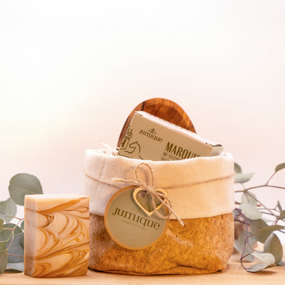 Ein Geschenkset steht auf einer Baumscheibe. In einem Korb aus Kork mit hellem Stoffrand ist mit einer braunen, geflochtenen Band ein grünes "Jumique" Schild und ein Herz aus Holz gebunden. In dem Korb befinden sich eine Seifenschale aus Olivenholz und die Kartonverpackung der Stutenmilchseife "Marquise" mit Hafer und Honig. Das Seifenstück steht aufrecht links neben dem Korb, sodass die goldene und braune Maserung sichtbar ist.