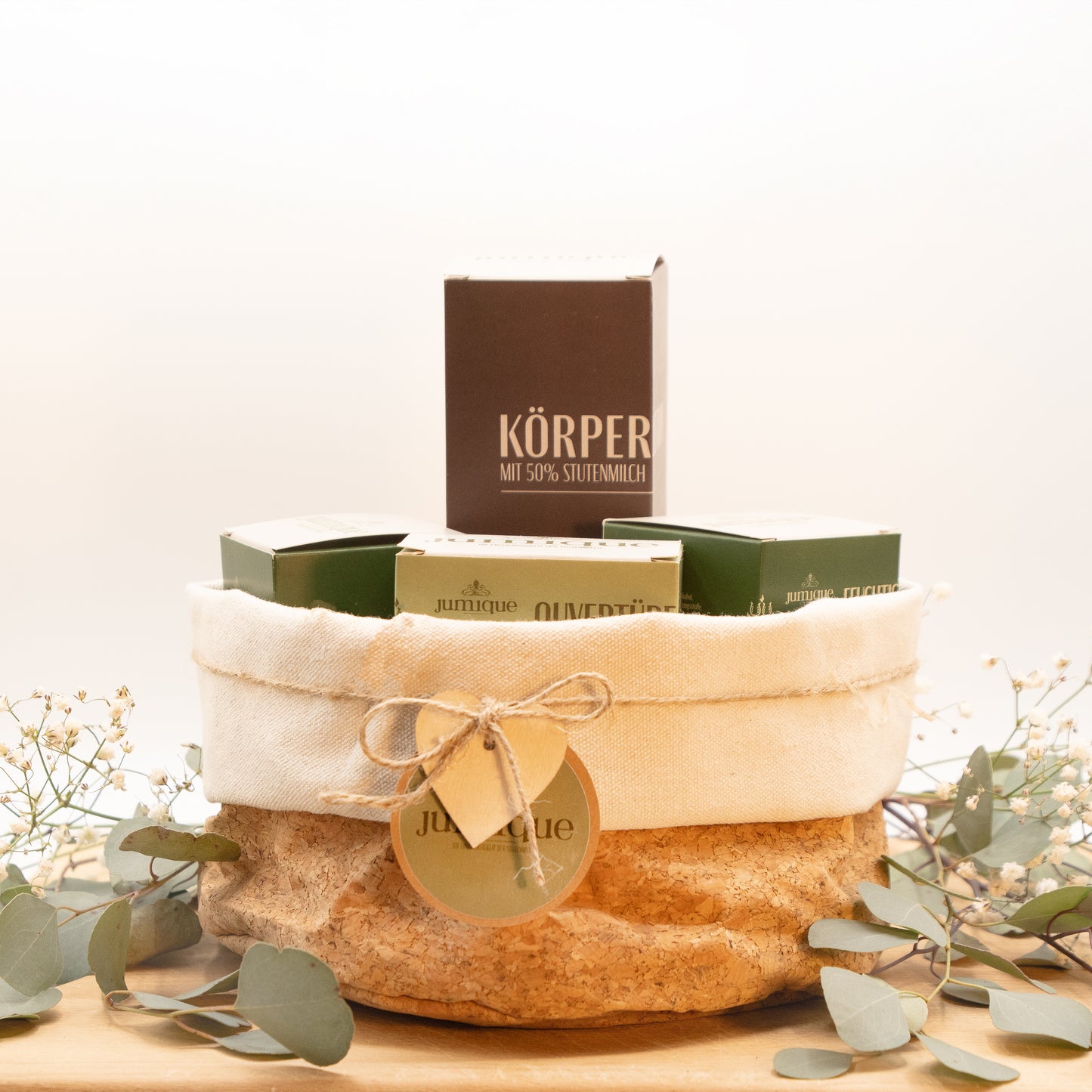 Ein Geschenkset steht auf einer Baumscheibe. In einem Korb aus Kork mit hellem Stoffrand ist mit einer braunen, geflochtenen Band ein grünes "Jumique" Schild und ein Herz aus Holz gebunden. In dem Korb befinden sich eingepackt in Kartonverpackungen die Stutenmilchseife "Ouvertüre" mit Kardamom und Pfeffer, eine Körperlotion, eine Aufbau- und eine Feuchtigkeitscreme. Das Jumique-Logo ist auf der Seifenverpackung zu sehen. Es zeigt zwei Pferde, die zu einem Wappen blicken, in dessen Mitte ein "H" steht.