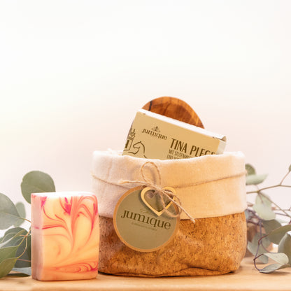 Ein Geschenkset steht auf einer Baumscheibe. In einem Korb aus Kork mit hellem Stoffrand ist mit einer braunen, geflochtenen Band ein grünes "Jumique" Schild und ein Herz aus Holz gebunden. In dem Korb befinden sich eine Seifenschale aus Olivenholz und die Kartonverpackung der Stutenmilchseife "Tina" mit Rosenduft. Das Seifenstück steht aufrecht links neben dem Korb, sodass die rosa Maserung sichtbar ist.