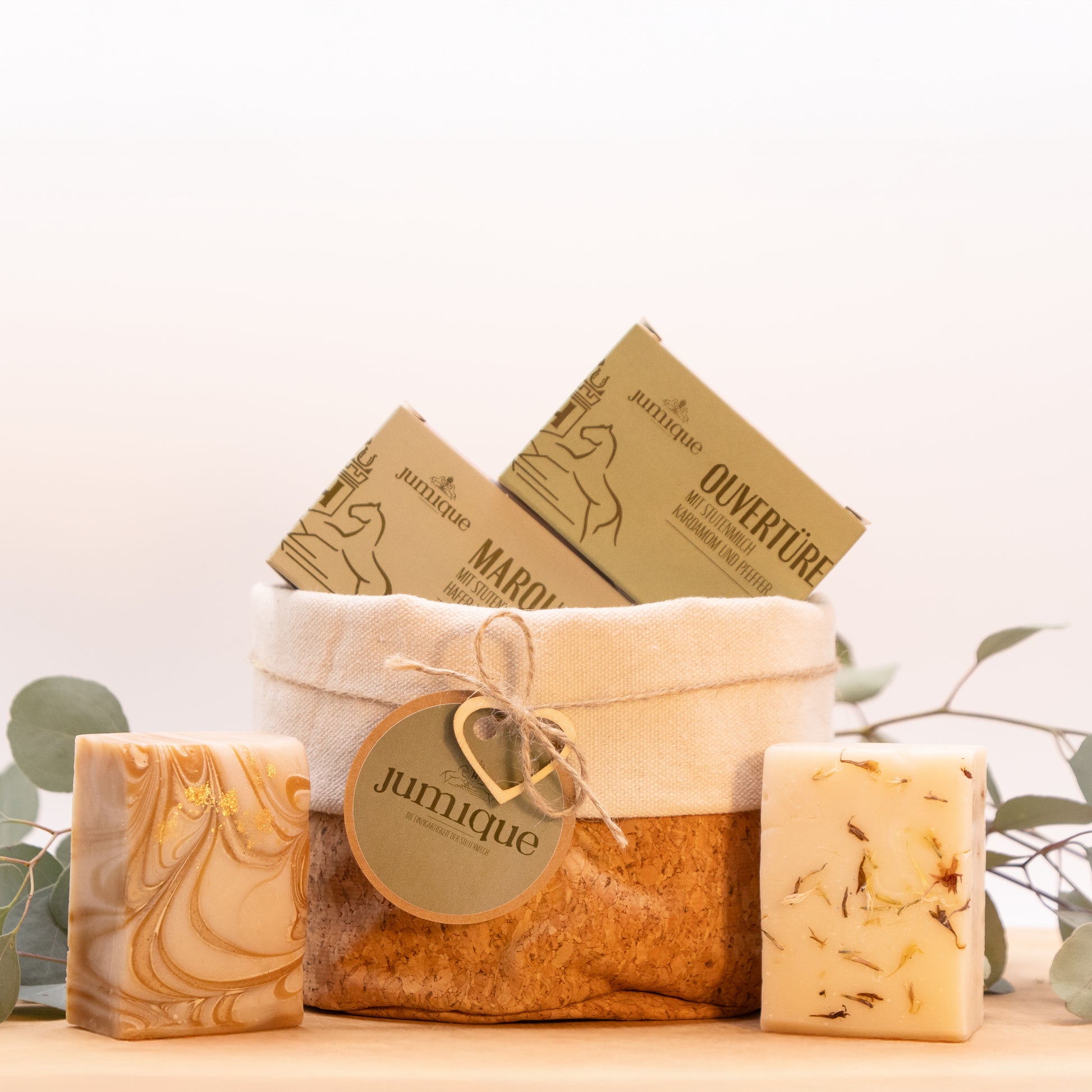 Ein Geschenkset steht auf einer Baumscheibe. In einem Korb aus Kork mit hellem Stoffrand ist mit einer braunen, geflochtenen Band ein grünes "Jumique" Schild und ein Herz aus Holz gebunden. In dem Korb befinden sich die Kartonverpackungen der Stutenmilchseifen "Marquise" mit Hafer und Honig und "Ouvertüre" mit Kardamom und Pfeffer. Das Seifenstücken stehen aufrecht rechts und links neben dem Korb.