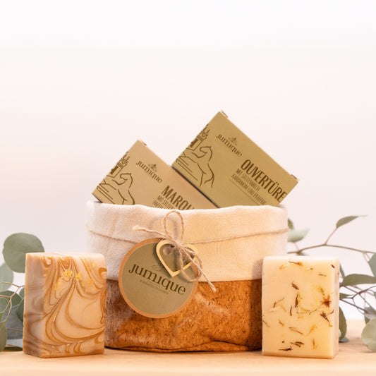 Ein Geschenkset steht auf einer Baumscheibe. In einem Korb aus Kork mit hellem Stoffrand ist mit einer braunen, geflochtenen Band ein grünes "Jumique" Schild und ein Herz aus Holz gebunden. In dem Korb befinden sich die Kartonverpackungen der Stutenmilchseifen "Marquise" mit Hafer und Honig und "Ouvertüre" mit Kardamom und Pfeffer. Das Seifenstücken stehen aufrecht rechts und links neben dem Korb.